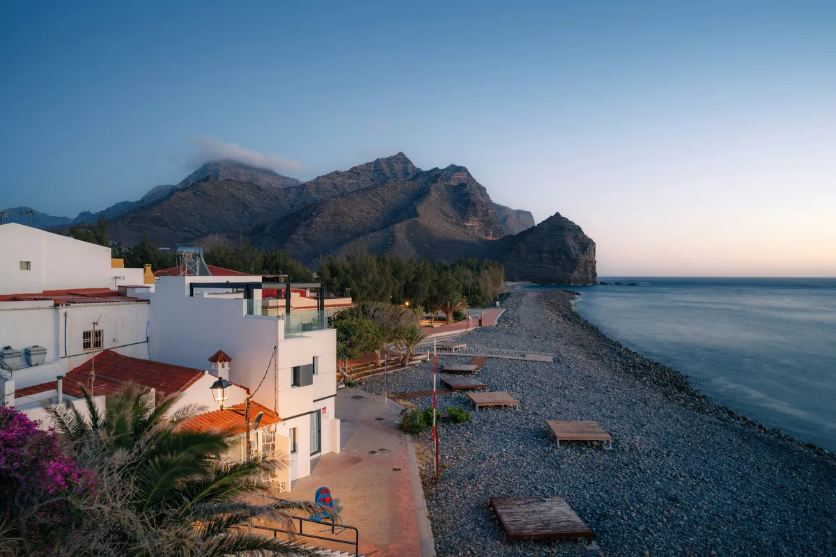 Playa de callaos de La Aldea al atardecer, con casas blancas y montañas junto al mar