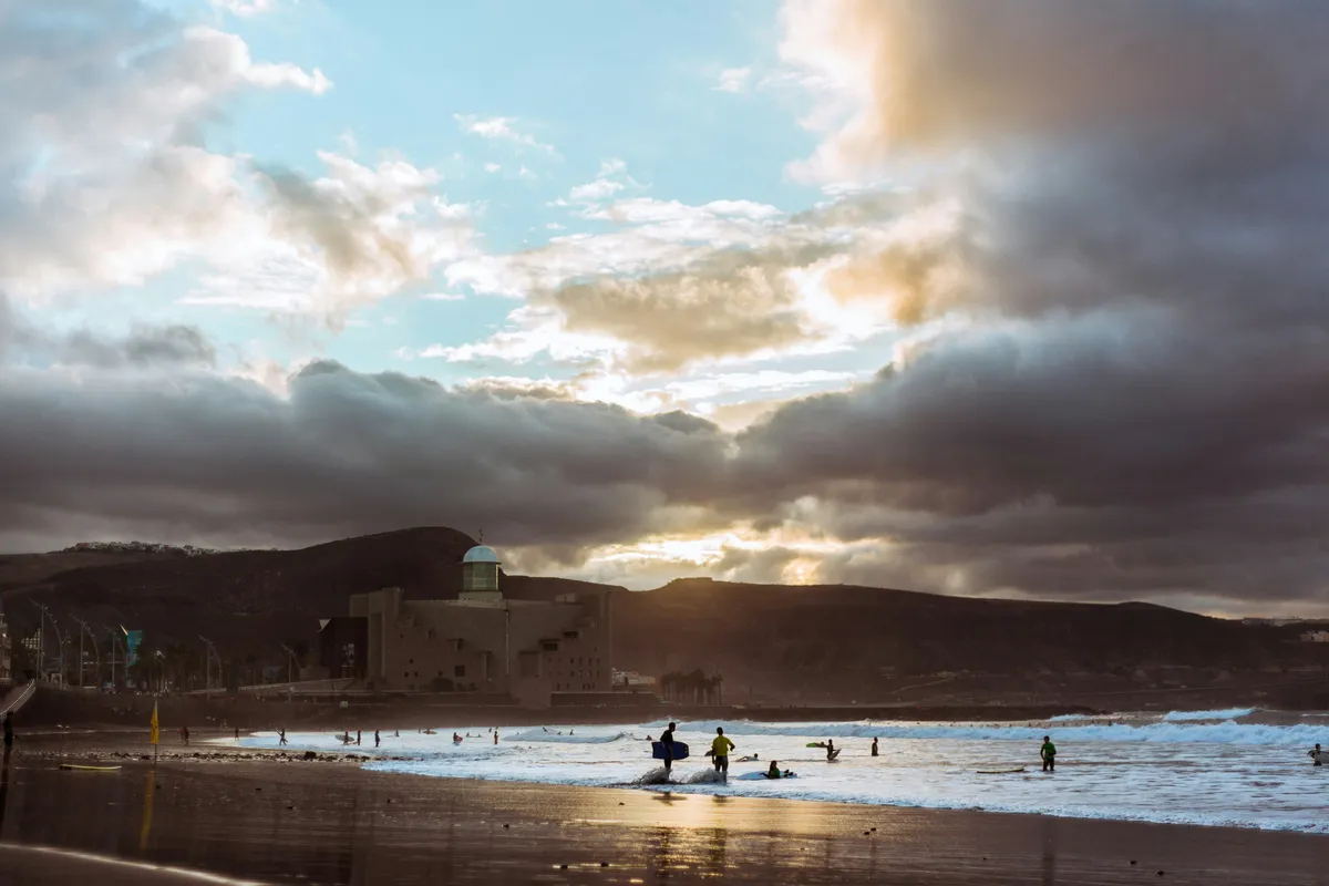 Surfistas en la orilla de Las Canteras al atardecer, con el Auditorio Alfredo Kraus al fondo