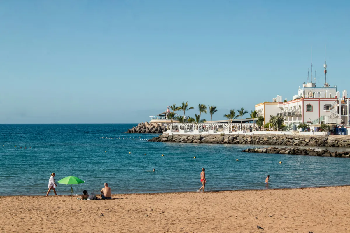 Bañistas en la playa de Mogán, junto al dique y los edificios blancos del puerto